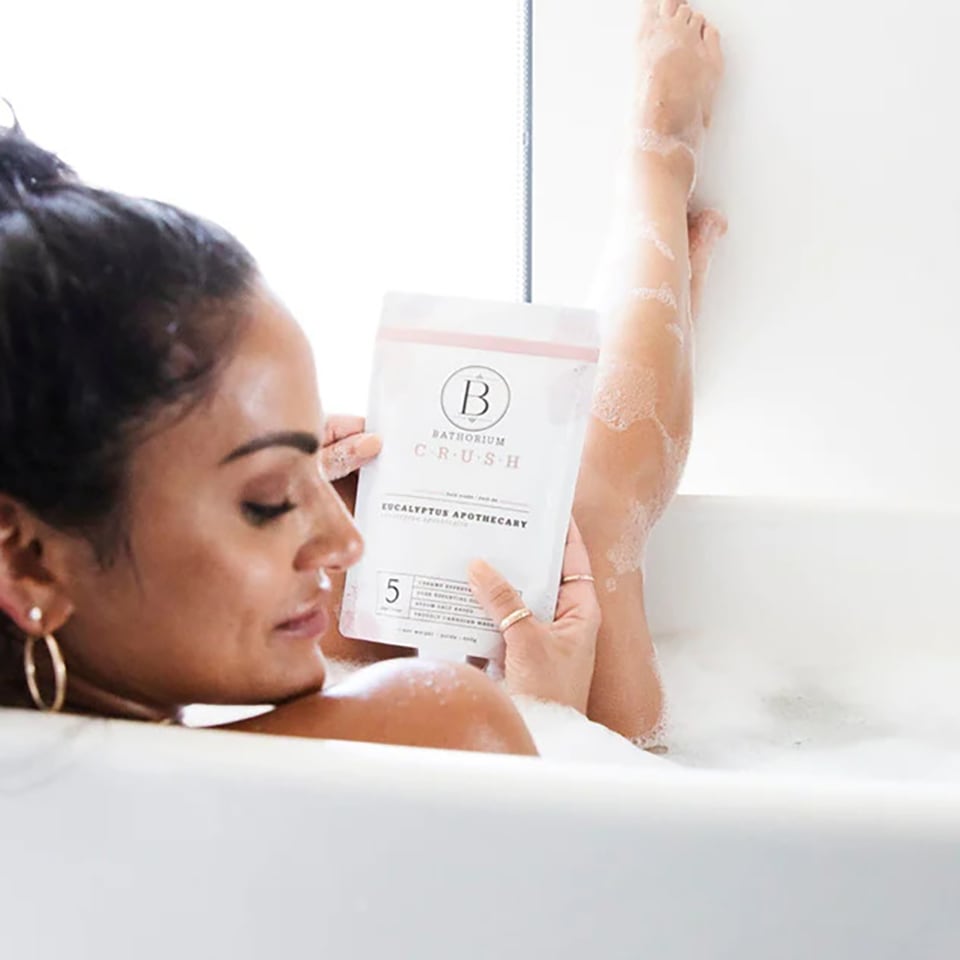Black woman in a bathtub, holding a package of Bathorium bath soaks, looking over her shoulder at the camera