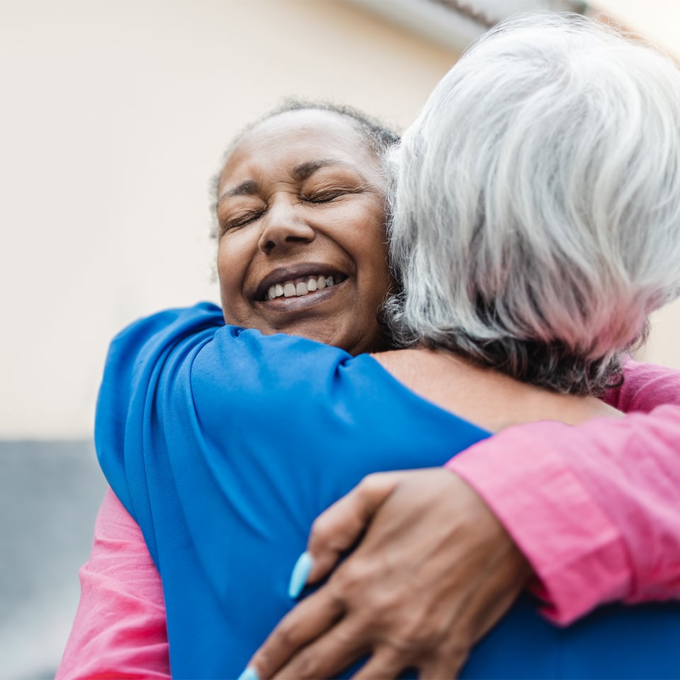 An elderly Black woman embraces a white woman with silver hair in a deep hug; her eyes are closed and she is facing the camera